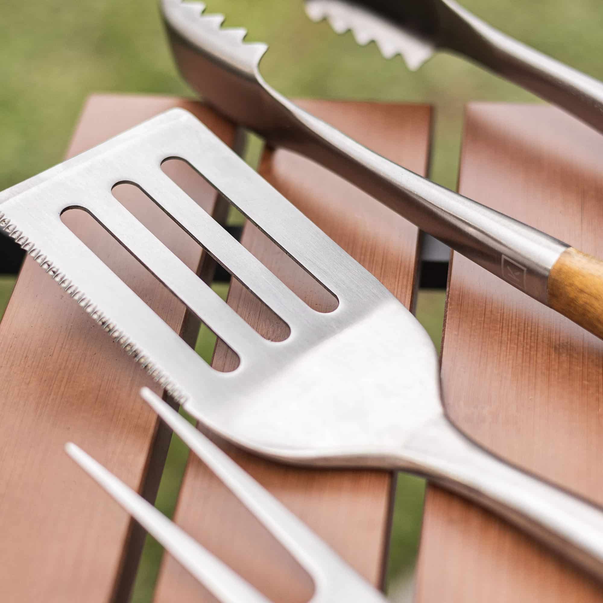 Ustensiles de barbecue en inox, dont une spatule dentelée, une pince et une fourchette, posés sur une tablette en bois.