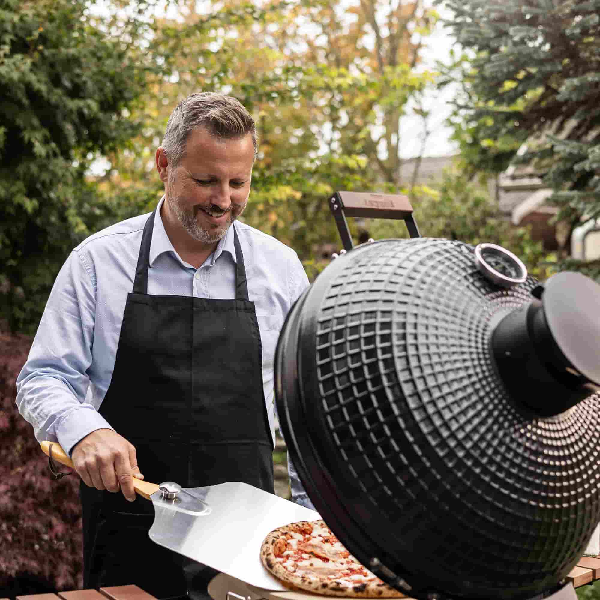 Homme retirant une pizza dorée du Kamado Forest Grill à l’aide d’une pelle à pizza, lors d’une cuisson en plein air.