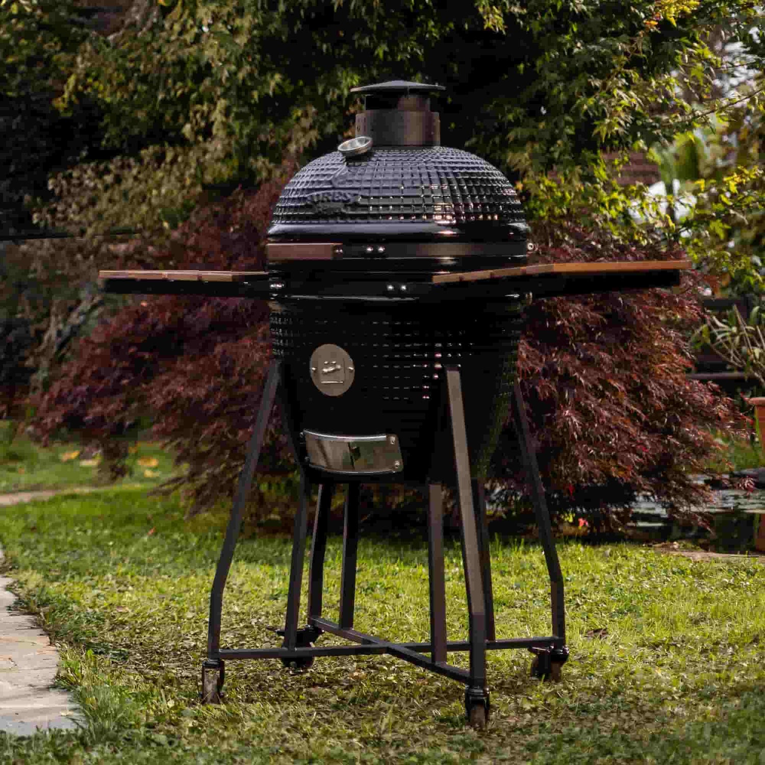 Barbecue Kamado Forest Grill en céramique noire avec tablettes latérales en bois, installé dans un jardin.