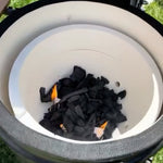 Vue intérieure d’un barbecue Kamado Forest Grill avec le foyer en céramique blanche, contenant du charbon de bois noir en train de s’enflammer légèrement, illustrant la phase d’allumage du barbecue.