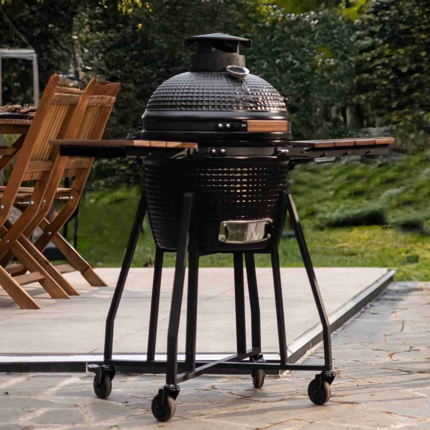 Barbecue Kamado Forest Grill en céramique noire avec tablettes latérales en bois, installé sur une terrasse à côté d’une table de jardin.