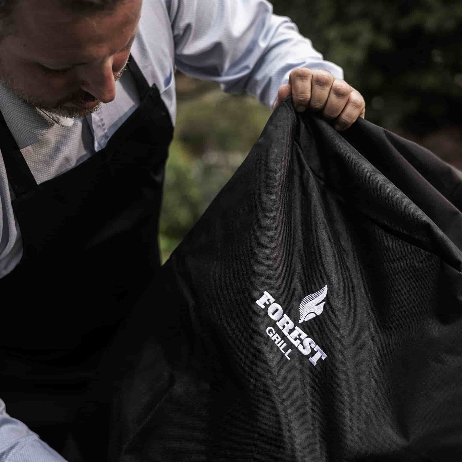 Homme tenant une housse noire de protection Forest Grill, conçue pour protéger le barbecue kamado des intempéries.