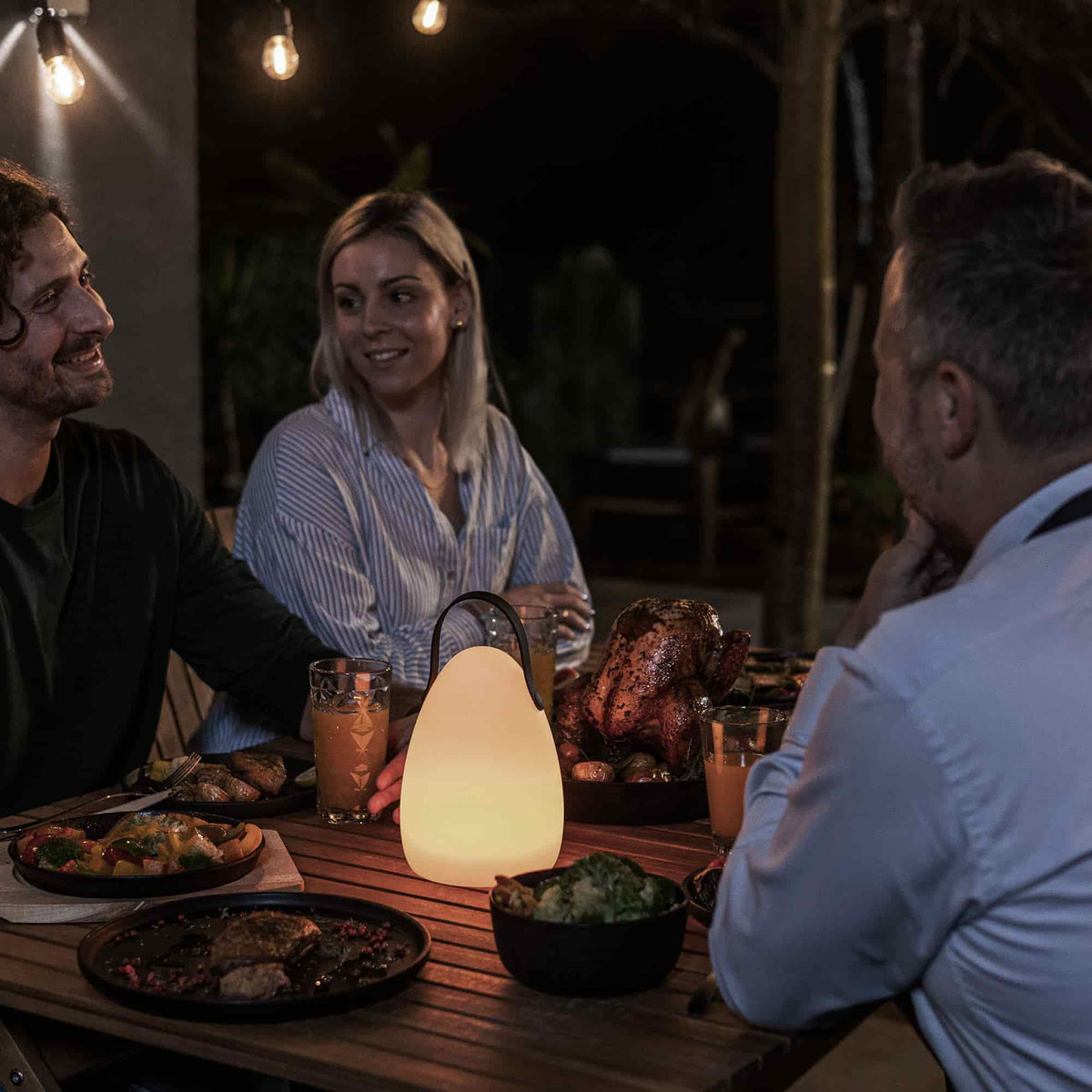 Trois personnes dînant en extérieur autour d’une table en bois, éclairée par une lampe LED en forme d’œuf posée au centre, créant une ambiance chaleureuse et conviviale.