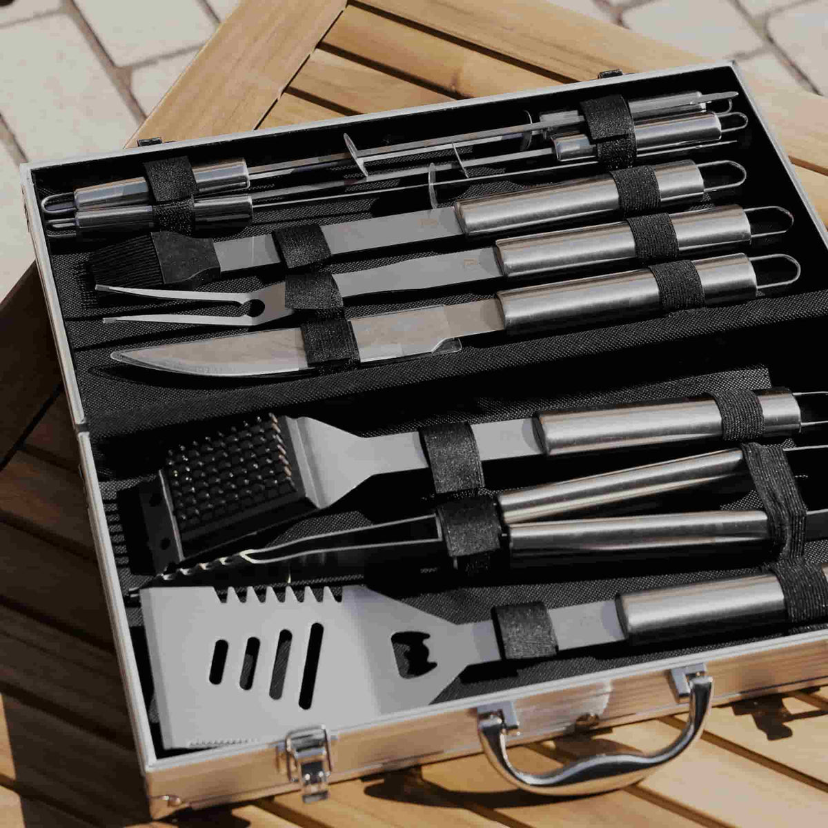 Mallette ouverte sur une table en bois, contenant un ensemble complet d’ustensiles de barbecue en acier inoxydable, rangés avec précision dans des sangles noires.
