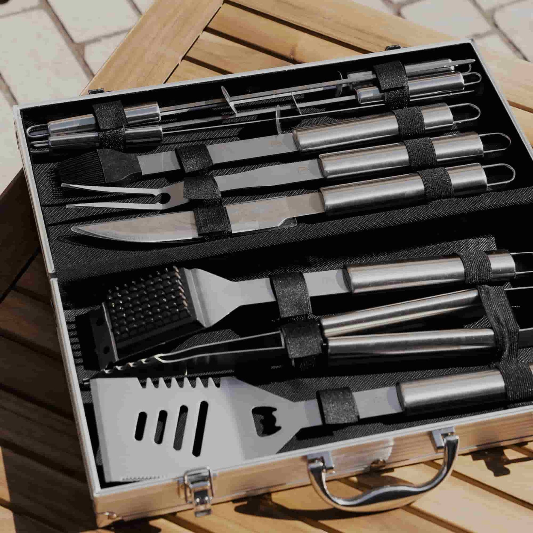 Mallette ouverte sur une table en bois, contenant un ensemble complet d’ustensiles de barbecue en acier inoxydable, rangés avec précision dans des sangles noires.