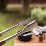 Gros plan sur une pince de barbecue en acier inoxydable avec manche en bois, tenue au-dessus d’une table d’extérieur.