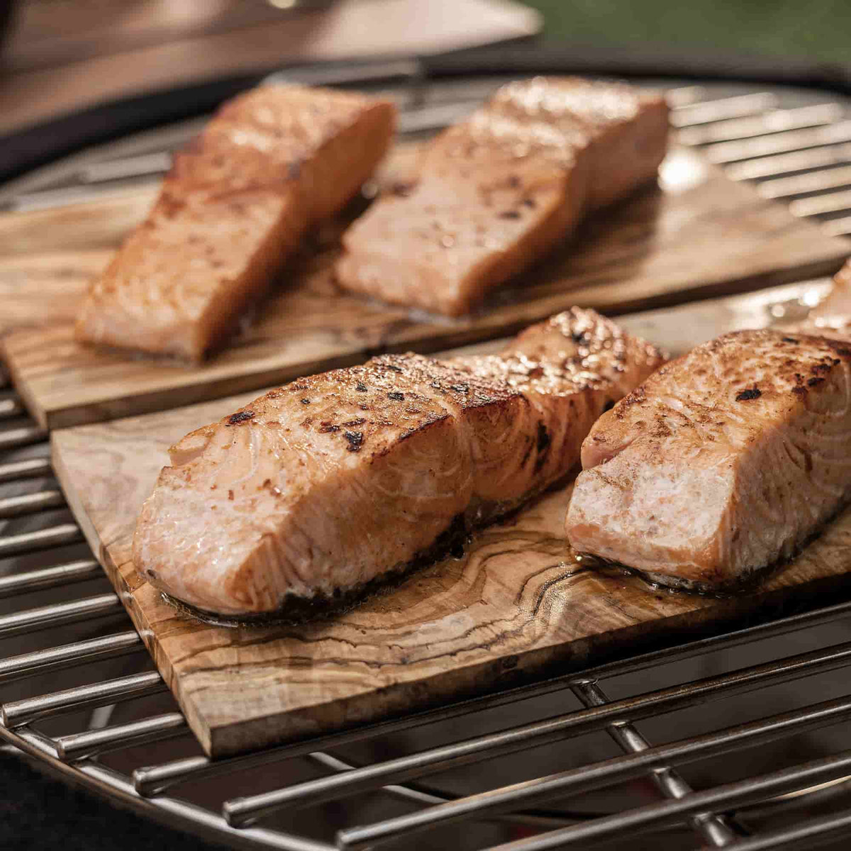 Filets de saumon grillés sur des planches en bois d’olivier, déposées sur la grille d’un barbecue kamado pour une cuisson parfumée et authentique.