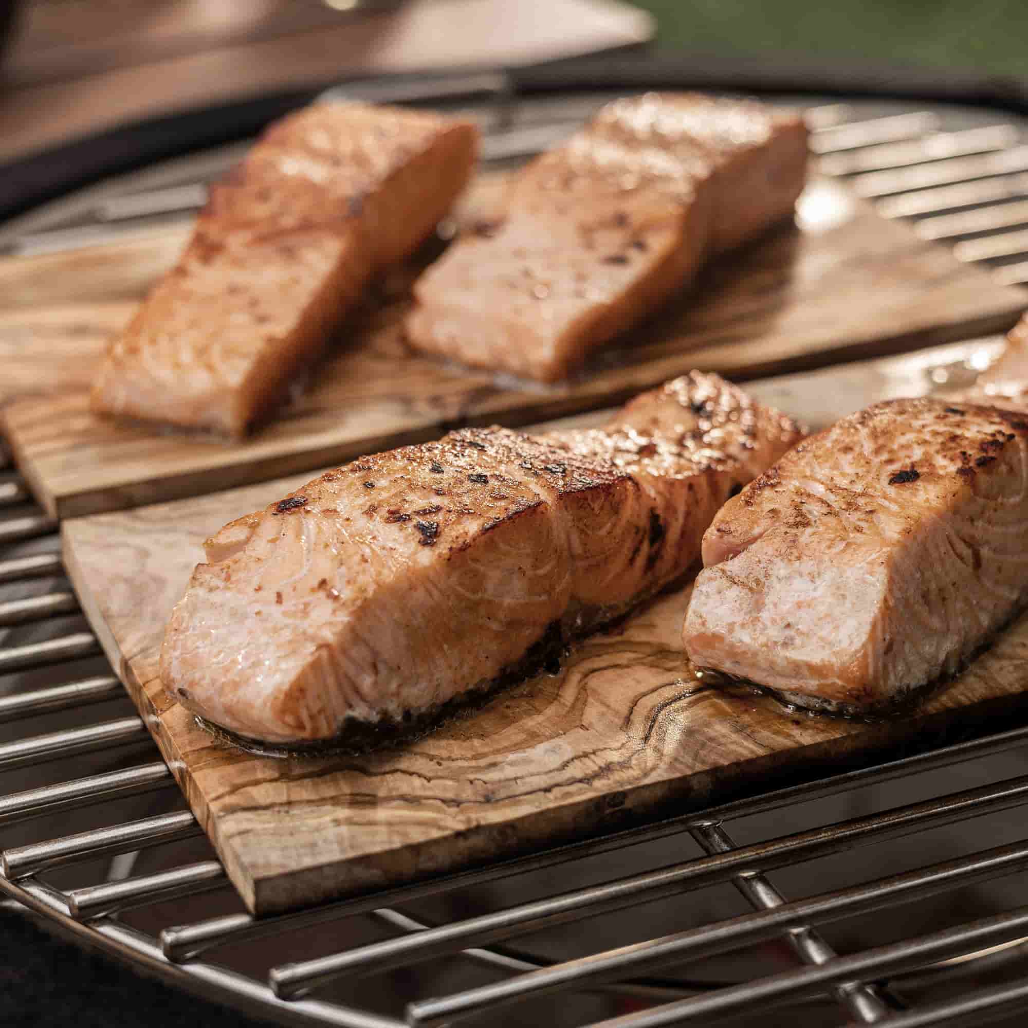 Filets de saumon grillés sur des planches en bois d’olivier, déposées sur la grille d’un barbecue kamado pour une cuisson parfumée et authentique.