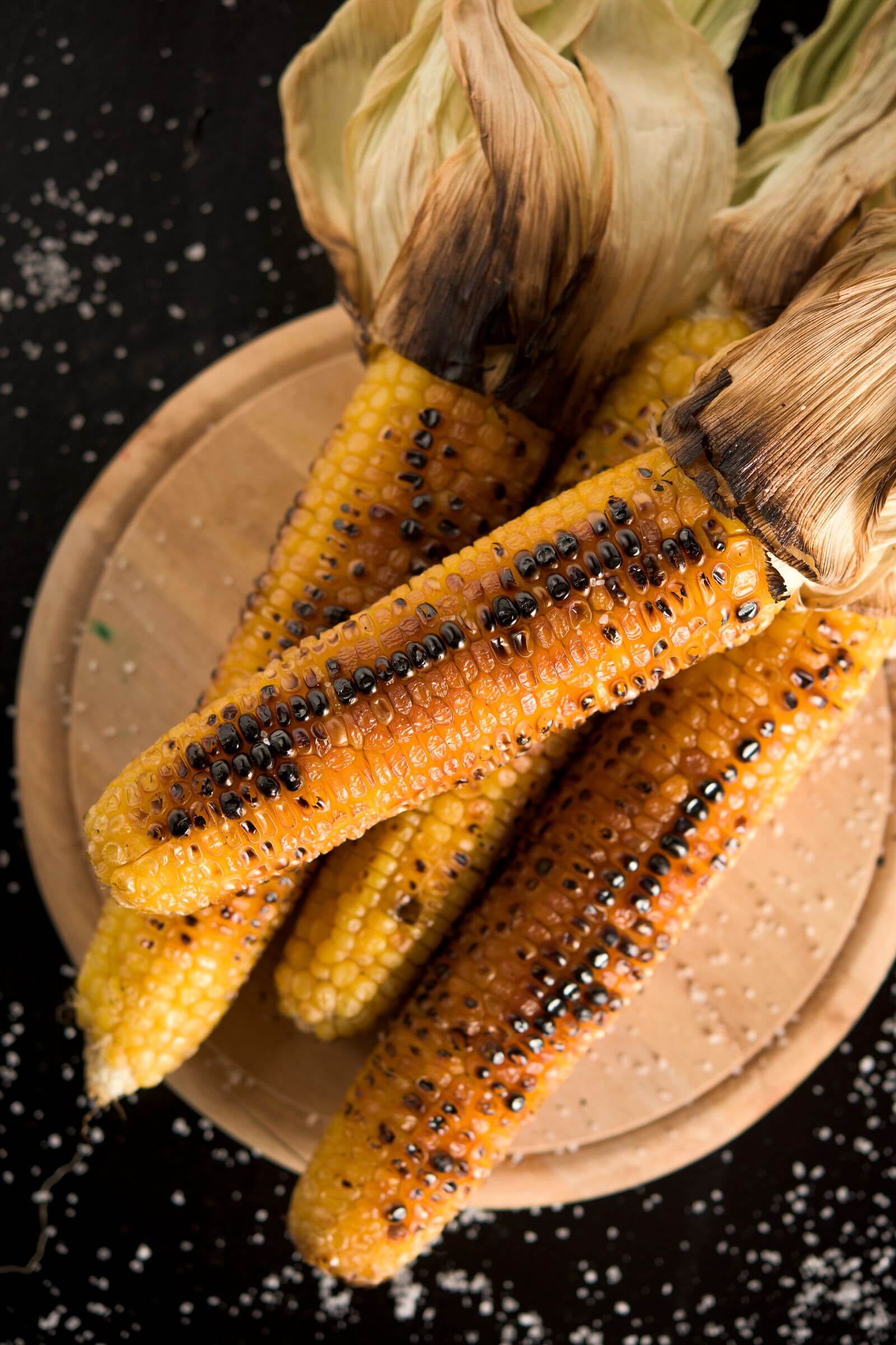 Épis de maïs grillés au kamado avec leurs feuilles, saupoudrés de gros sel, posés sur une planche en bois.