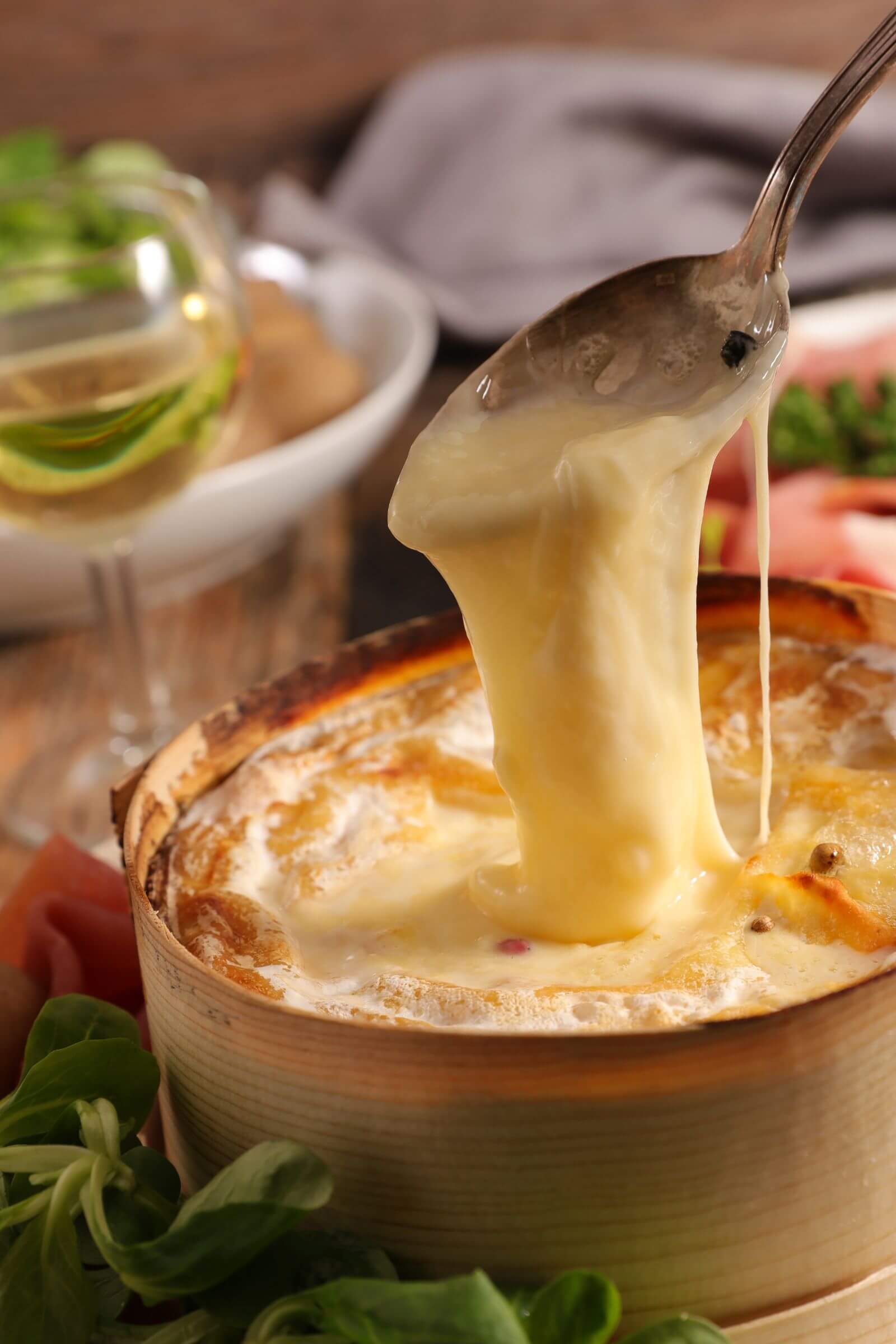 Fromage Mont-d’Or chaud et fondant dans sa boîte en bois, servi avec une cuillère et accompagné de salade et vin blanc.