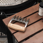 Griffe à viande en acier inoxydable avec manche en bois, posée sur une tablette de barbecue à côté d’un Kamado et de pots d’épices.