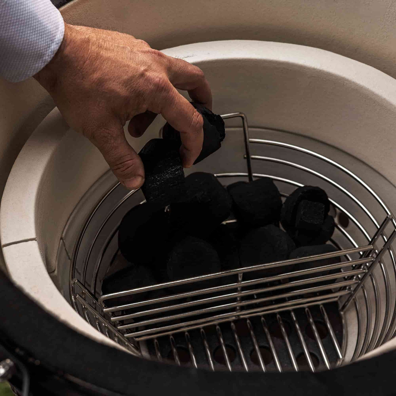 Main plaçant des morceaux de charbon dans un panier en acier inoxydable à l’intérieur d’un barbecue Kamado, avant l’allumage pour la cuisson.