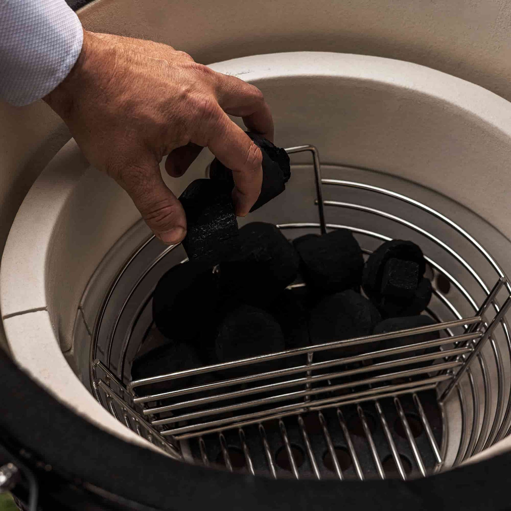 Main plaçant des morceaux de charbon dans un panier en acier inoxydable à l’intérieur d’un barbecue Kamado, avant l’allumage pour la cuisson.