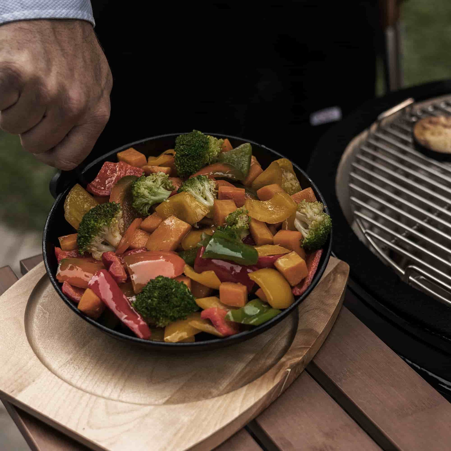 Main tenant un poêlon en fonte rempli de légumes colorés grillés, posé sur un dessous en bois à côté d’un barbecue Kamado, prêt à être servi.