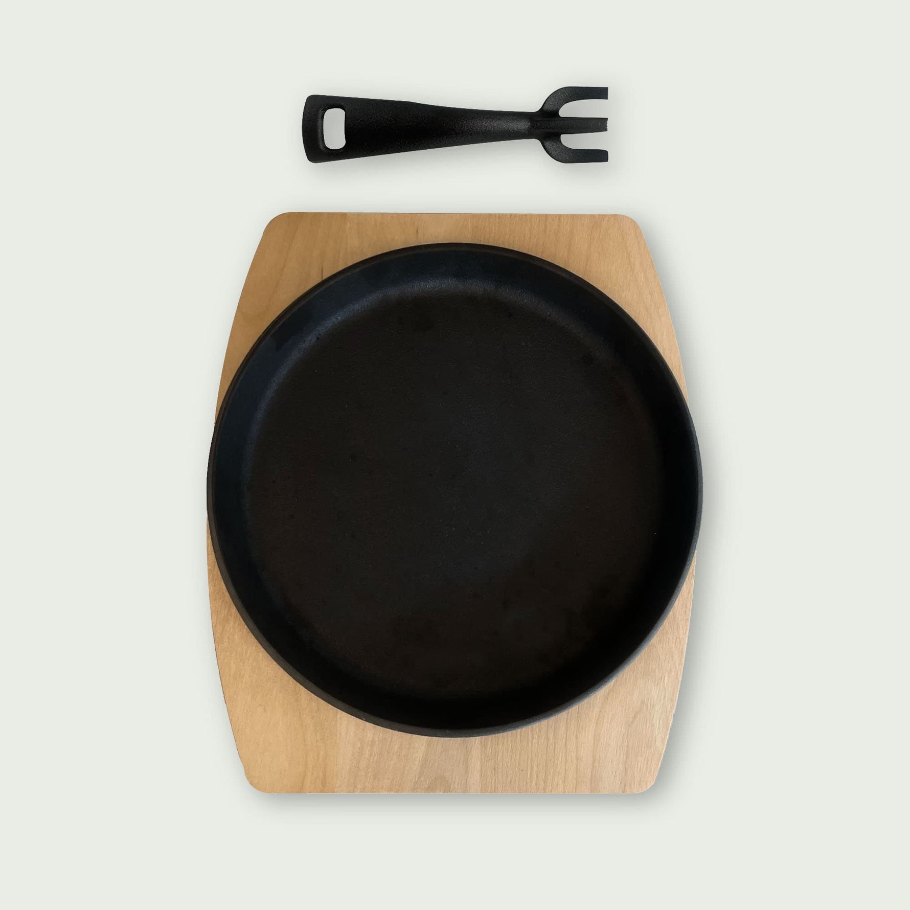Poêlon rond en fonte noire posé sur un dessous en bois clair, accompagné d’une poignée amovible noire placée au-dessus, idéal pour servir directement les plats chauds à table.