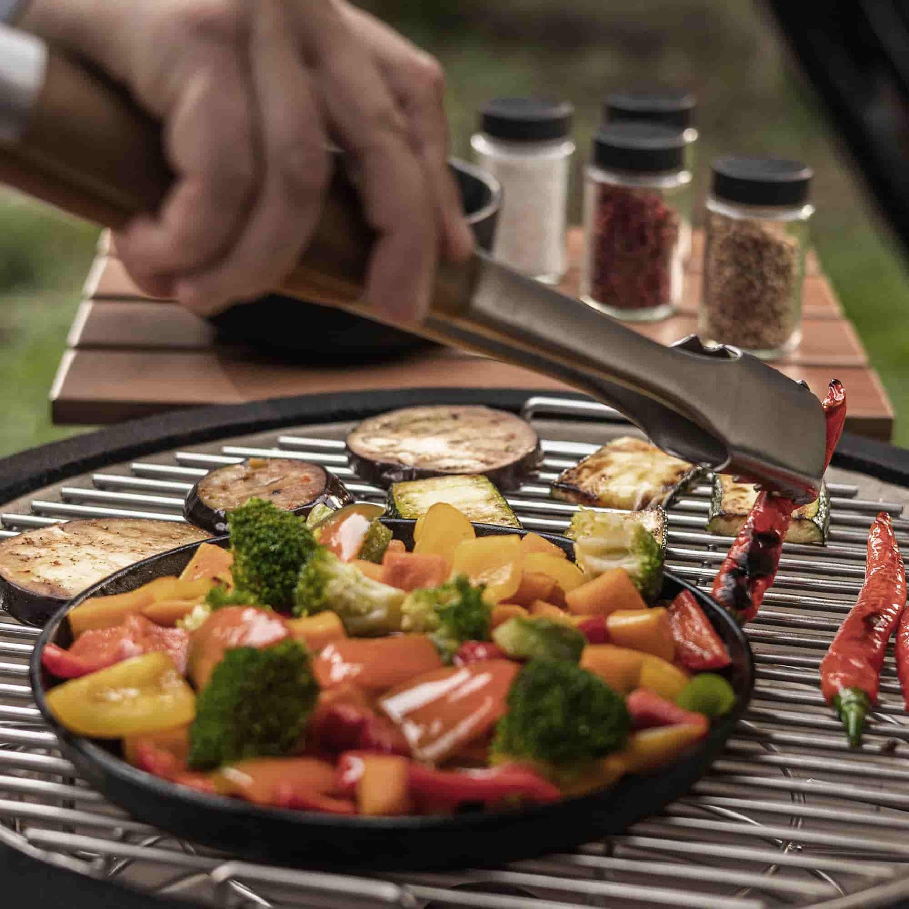 Légumes colorés en train de griller sur un Kamado, disposés dans une poêle en fonte noire ; une main utilise une pince de cuisson en inox pour retourner les aliments, avec des épices visibles en arrière-plan.