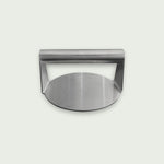 Presse à burger en acier inoxydable avec poignée intégrée, présentée sur fond clair, de face, idéale pour aplatir et cuire uniformément les steaks sur plancha ou barbecue.