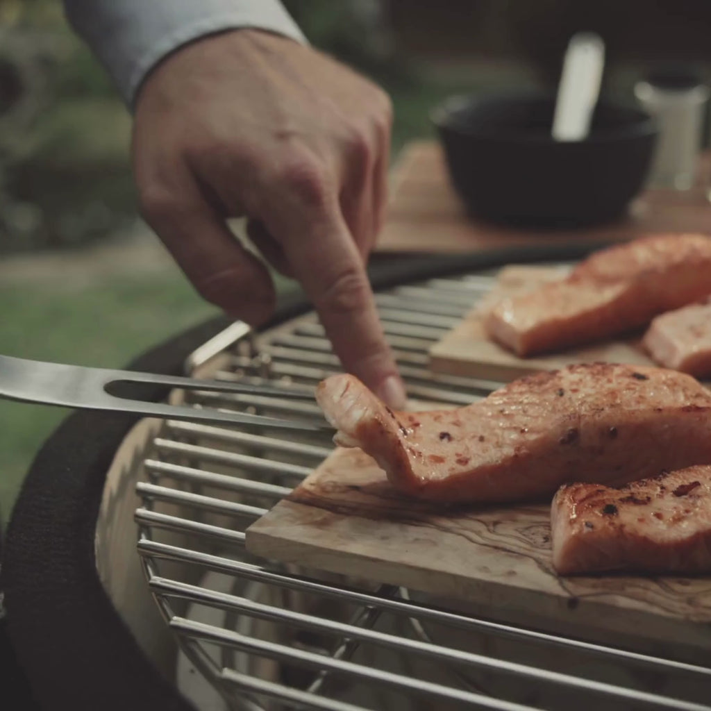 Vidéo présentant une planche de fumage en bois d’olivier, montrée en gros plan puis utilisée pour diffuser son arôme lors de la cuisson. On y voit la planche se consumer légèrement pour parfumer les aliments.
