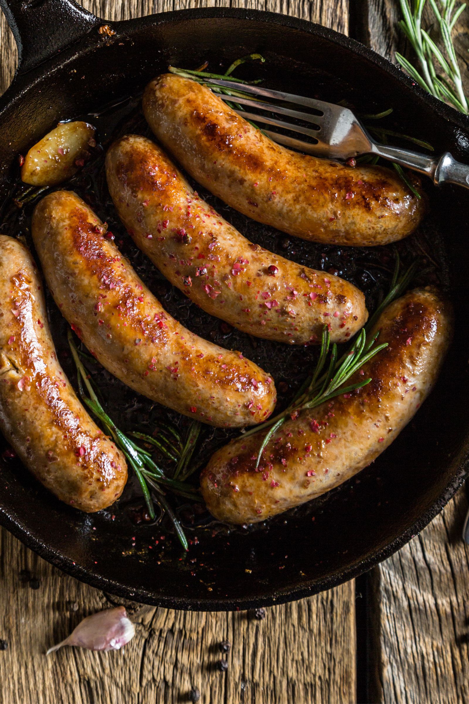 Saucisses dorées et grillées servies dans une poêle en fonte avec des herbes et des grains de poivre.