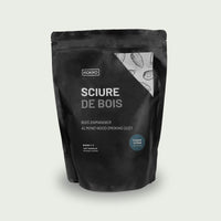 Sac de sciure de bois d’amandier posé sur une table en bois près d’un barbecue, accompagné d’une pince de cuisson en métal et bois au premier plan, dans un jardin.