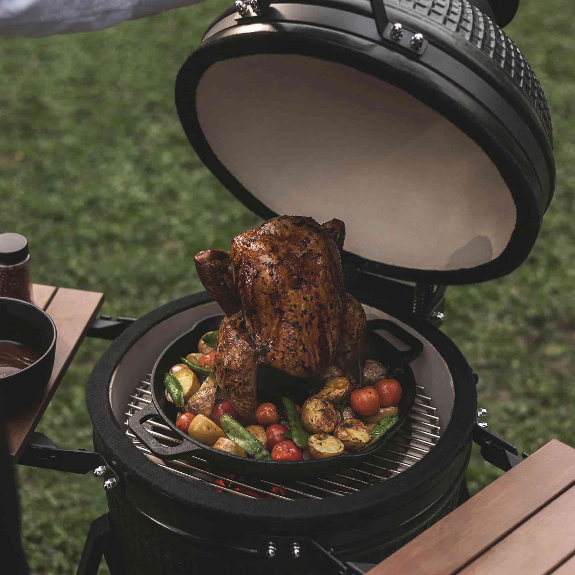 Poulet rôti placé verticalement sur un support de cuisson en fonte à l’intérieur d’un Kamado, entouré de légumes grillés colorés sur une grille au-dessus des braises.