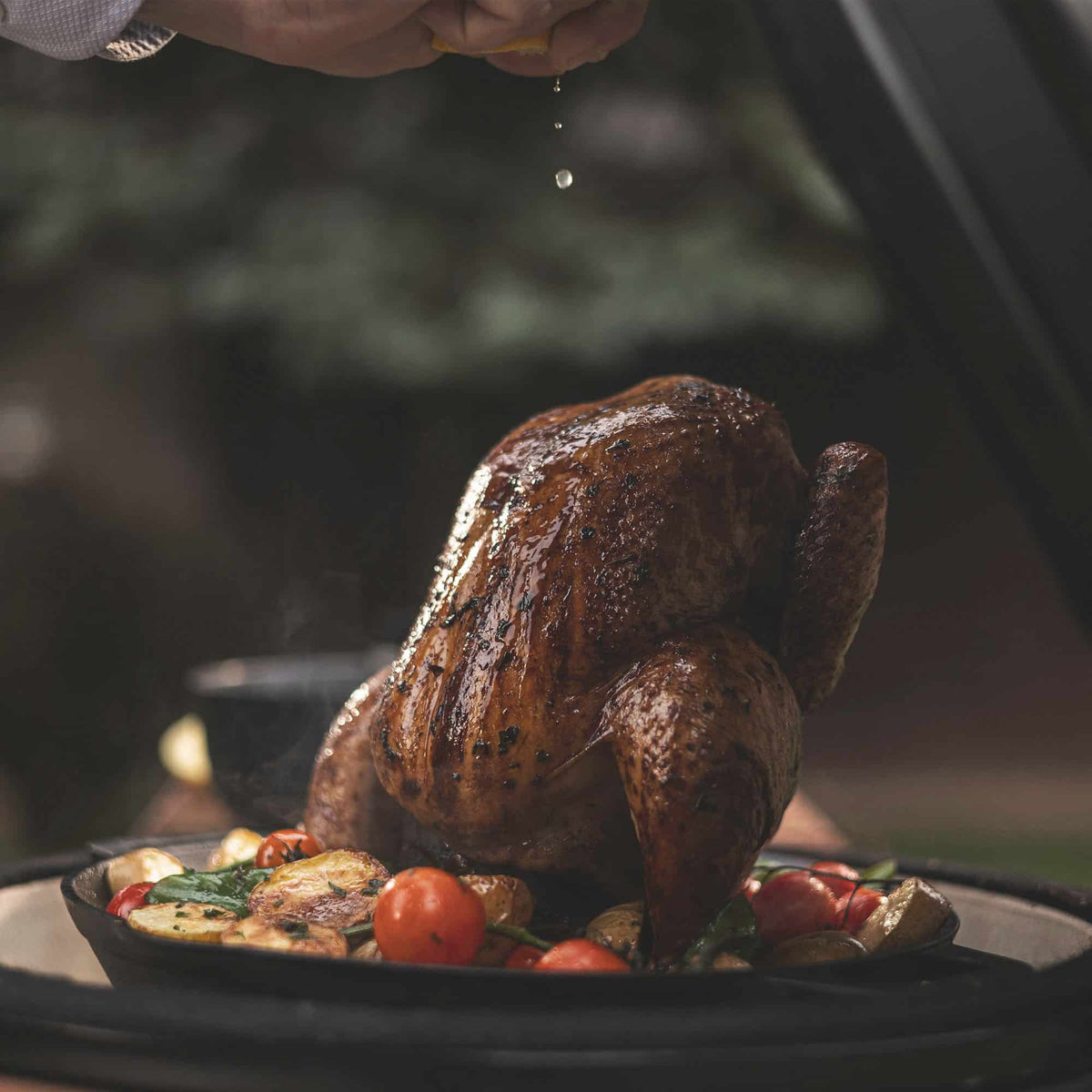 Gros plan sur un poulet rôti doré et juteux posé sur un support de cuisson en fonte, entouré de légumes grillés, tandis qu’une main presse un citron au-dessus pour ajouter du jus frais.
