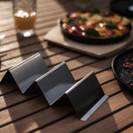 Support à tacos en inox brossé sur une table en bois, prêt à accueillir de délicieux tacos faits maison, avec des plats colorés en arrière-plan.