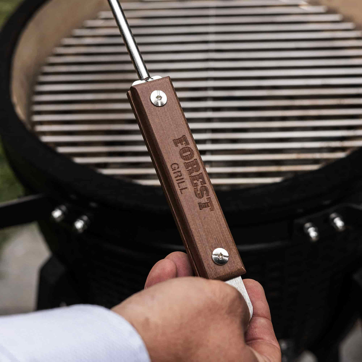 Main tenant un tire-cendre en acier inoxydable avec manche en bois gravé “Forest Grill”, devant un barbecue Kamado ouvert prêt à être nettoyé.