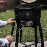 Personne utilisant un tire-cendre en acier inoxydable pour nettoyer la trappe inférieure d’un barbecue Kamado noir, posé sur son support métallique en extérieur.