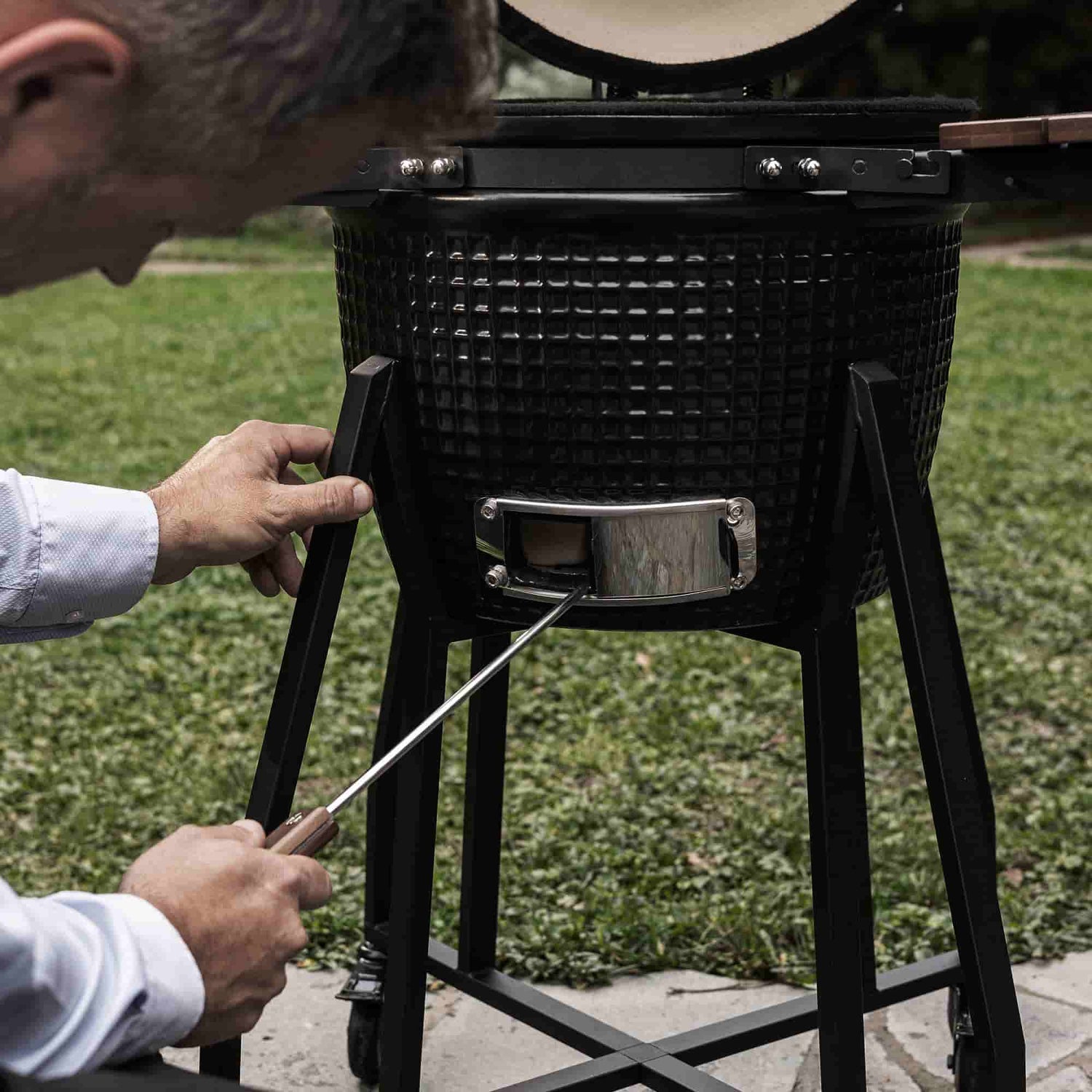Personne utilisant un tire-cendre en acier inoxydable pour nettoyer la trappe inférieure d’un barbecue Kamado noir, posé sur son support métallique en extérieur.