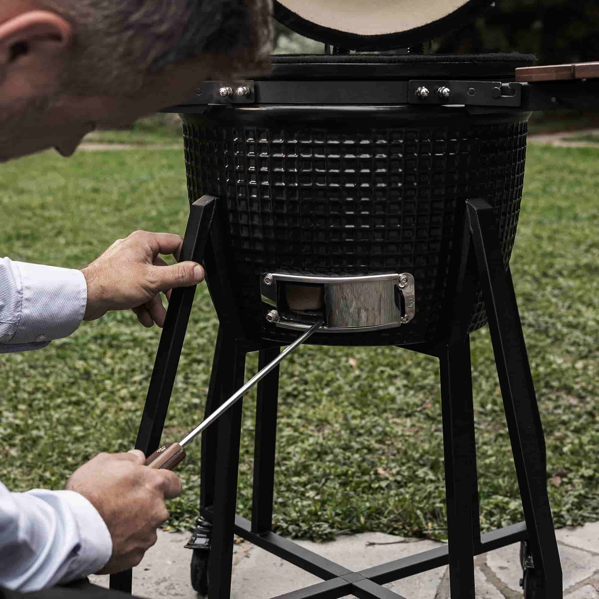Personne utilisant un tire-cendre en acier inoxydable pour nettoyer la trappe inférieure d’un barbecue Kamado noir, posé sur son support métallique en extérieur.